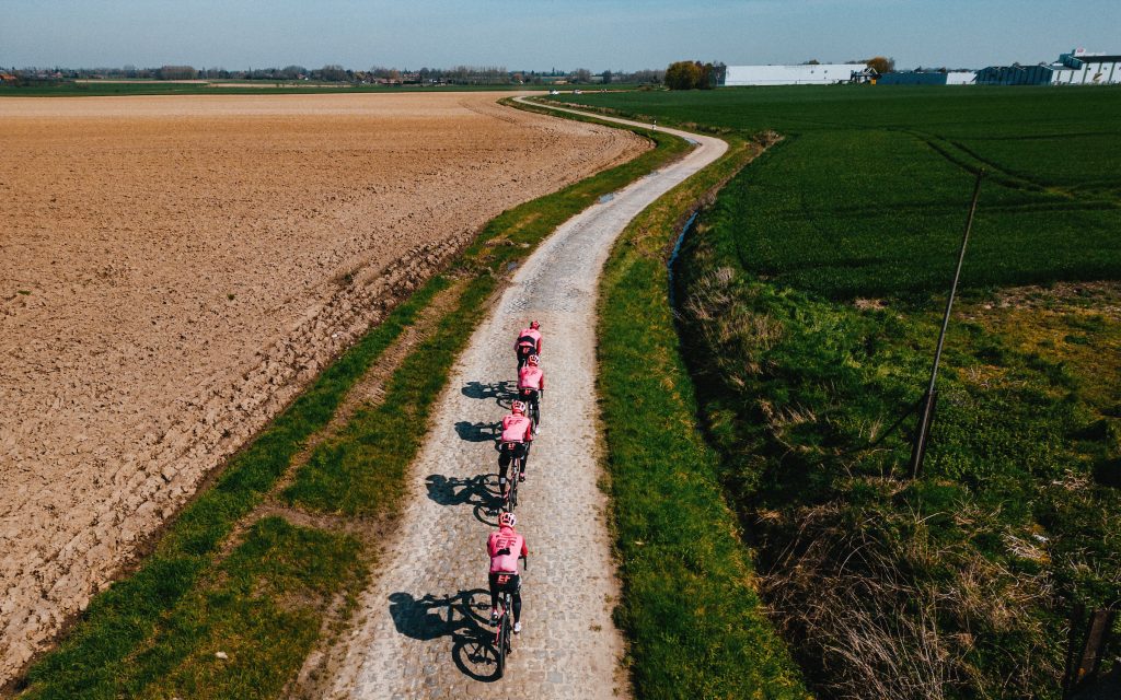 EF riders navigate another sector of cobbles. The overhead shot shows the riders going away from the camera down a narrow cobbled lane that snakes away in the distance. To the right, a green field and ditch, while to the left, the stark brown of freshly plowed ground contrasts.