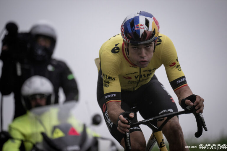 Wout van Aert chases alone at E3 Prijs after crashing. He's tailed by a TV moto and is looking up the road with his cheeks blowing out from the effort.